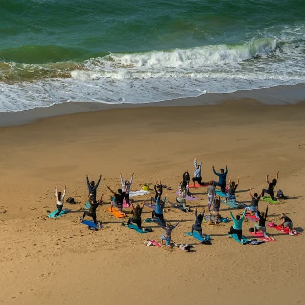 In-Person Groups - Photo of a group on retreat doing a yoga session led by Rose Long on a beach on a sunny day, facing the sea.
