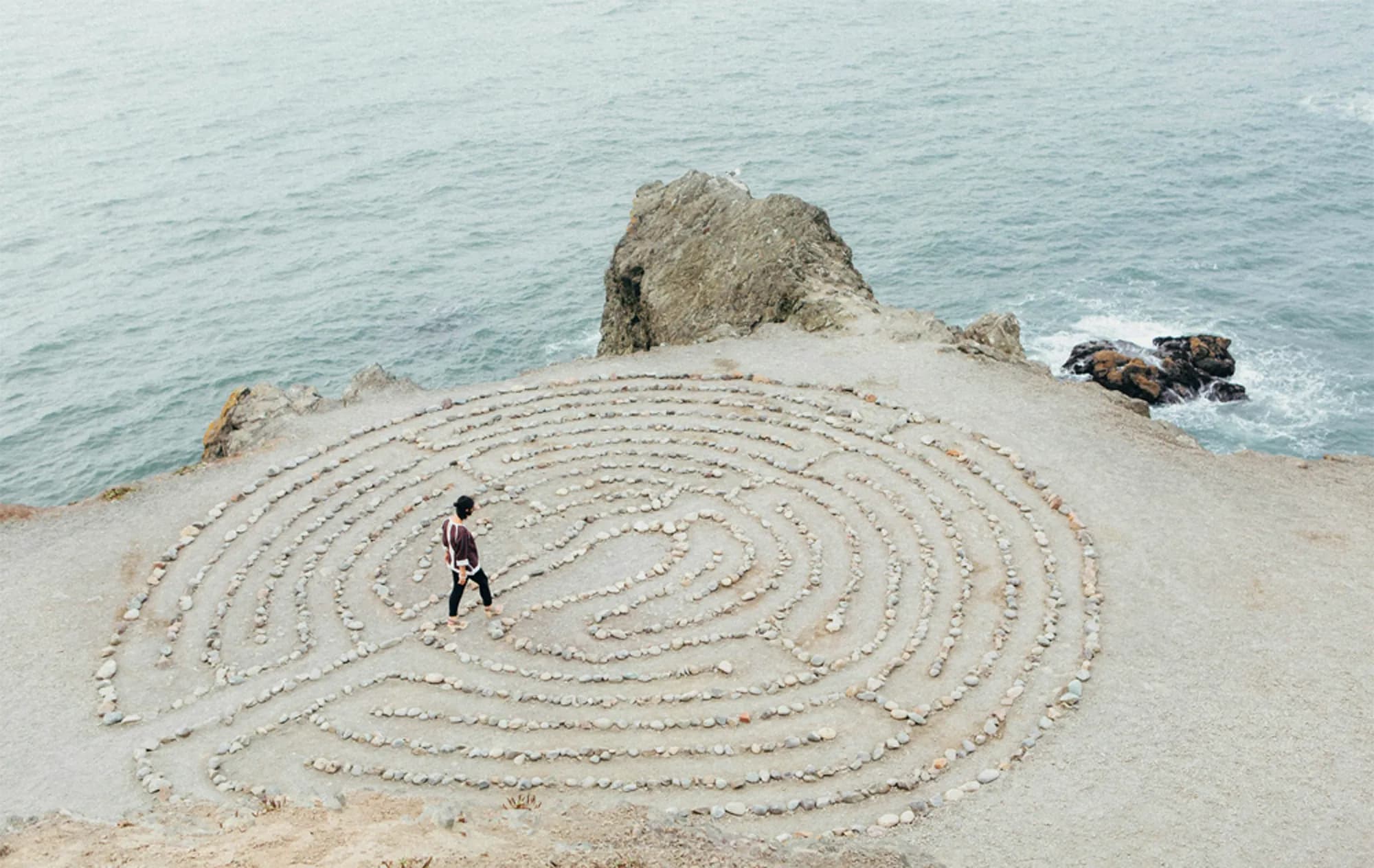 Person walking through a round pebble maze on a sandy beach on a clear day.