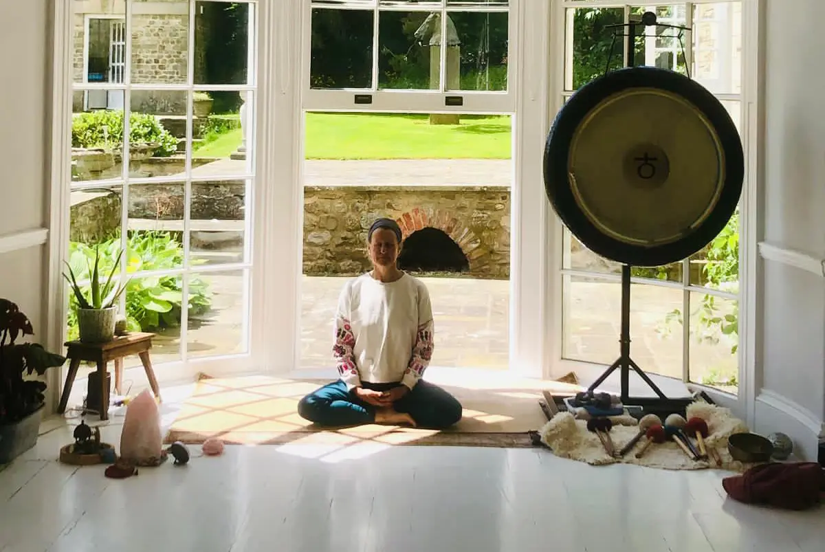Rose Long sat leading a retreat class in cross-legged position on the floor, smiling with her eyes closed. She is sat in-front of a large bay window which looks out onto a large sun-filled garden. There is a large, golden gong to her left and a plant with a himilayan salt lamp on her right.